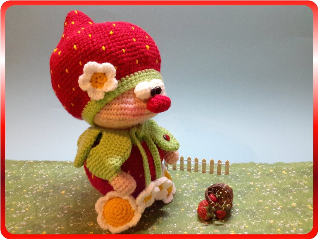 Gehäkelte Erdbeer-Amigurumi mit rotem Hut, Blüte und kleinem Korb auf grüner Miniwiese