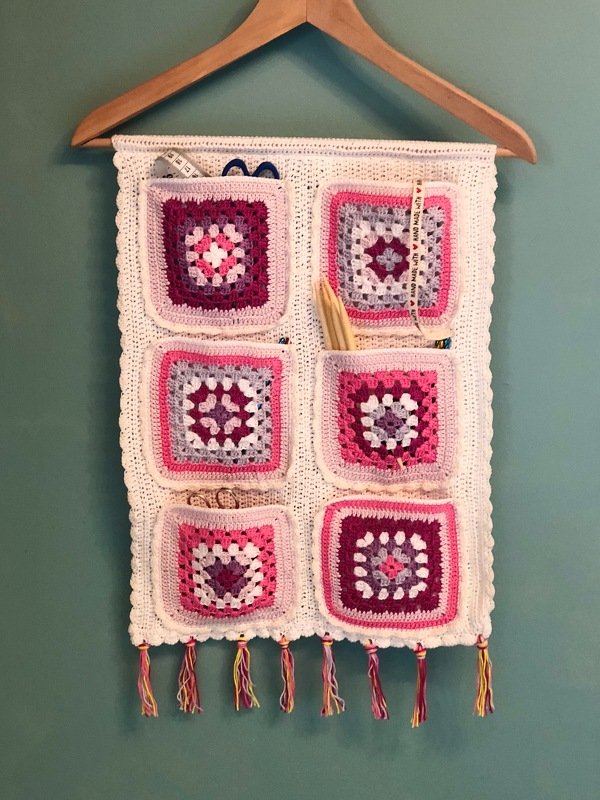 Gehäkeltes Wandutensilo mit sechs Granny‑Square‑Taschen, an einem Holzbügel hängend, mit Quasten.