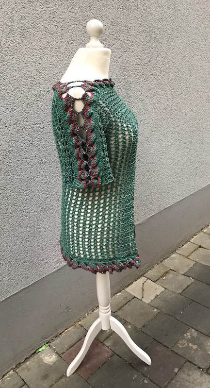 Grünes gehäkeltes Damenshirt mit kurzen Ärmeln und Schnürdetails am Ausschnitt und an den Ärmeln auf einer Schneiderpuppe vor grauer Wand.
