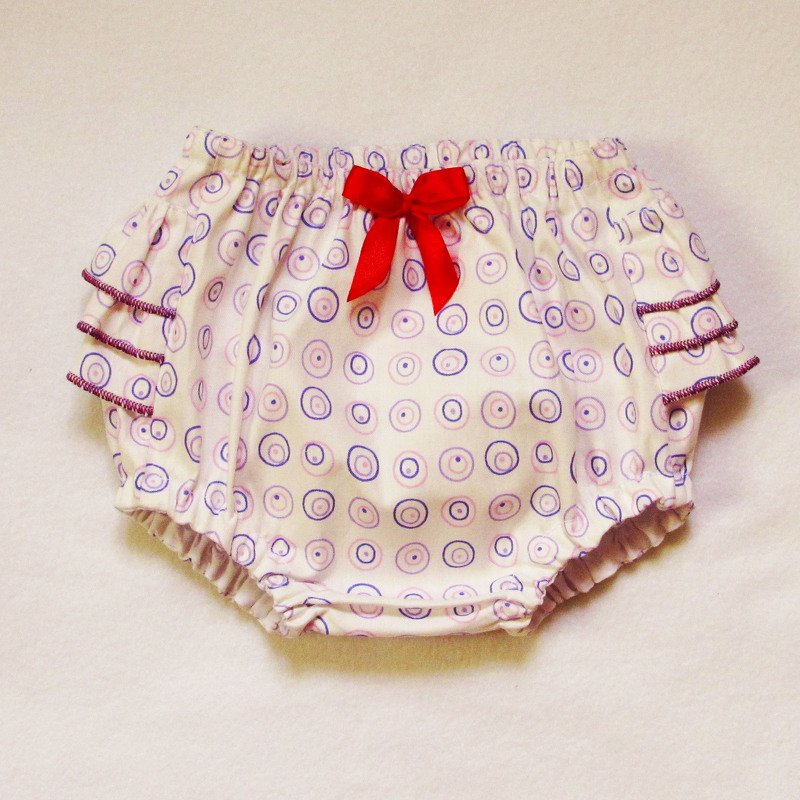 White baby bloomers with purple circle print, ruffled legs, and red bow