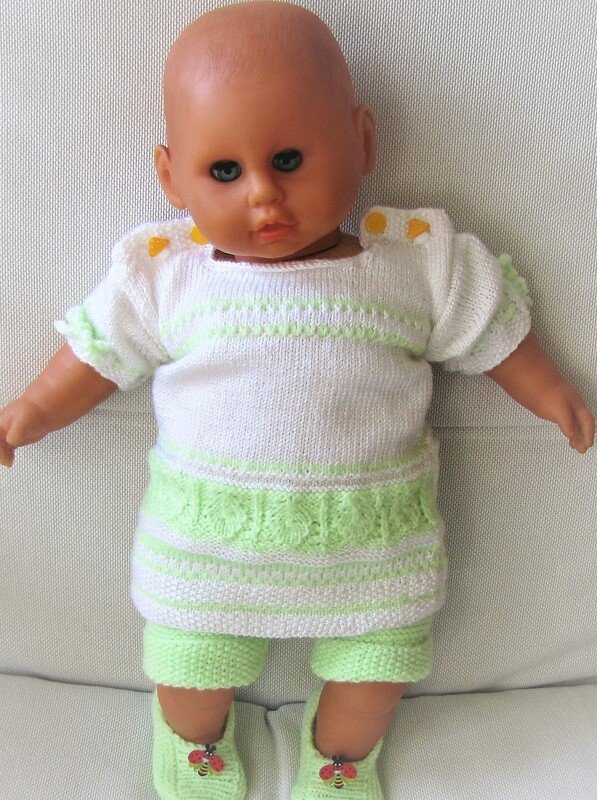 Babypuppe trägt ein gestricktes weiß-hellgrünes Set mit Oberteil, kurzer Hose und Schühchen mit Marienkäfern.
