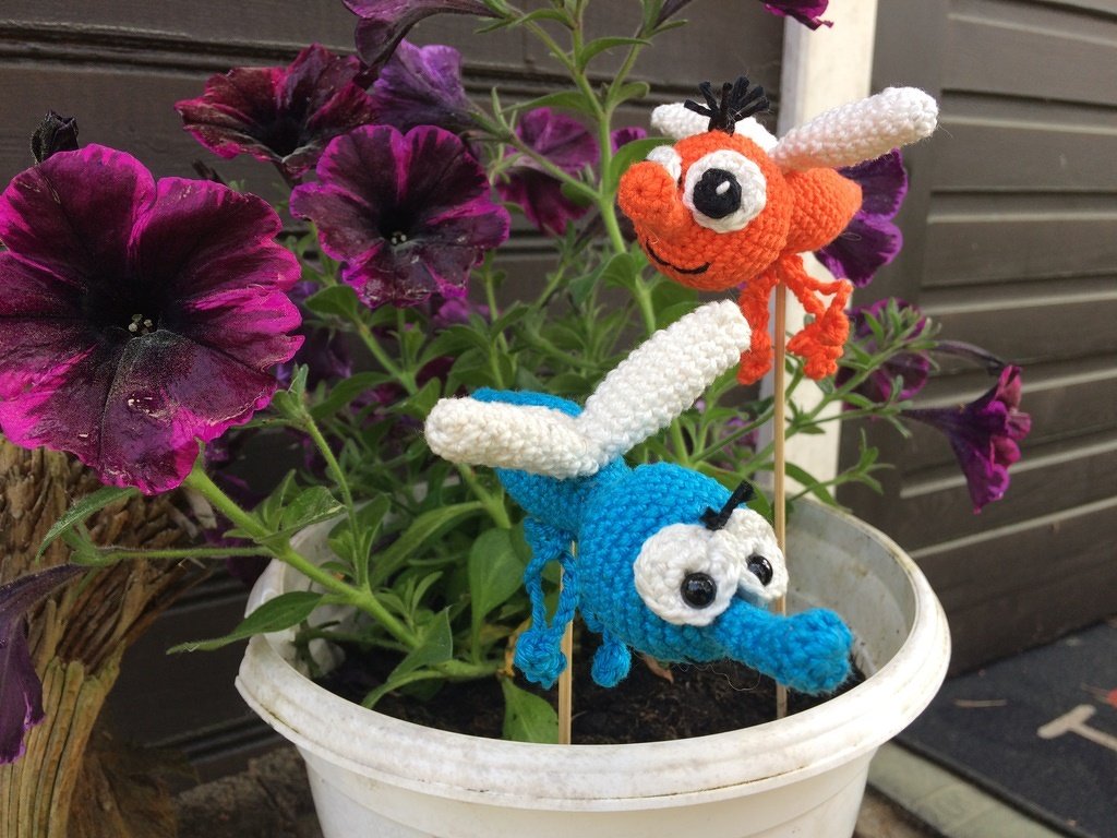 Zwei gehäkelte Insekten-Amigurumi (orange und blau) als Pflanzenstecker in einem weißen Blumentopf mit violetten Petunien.