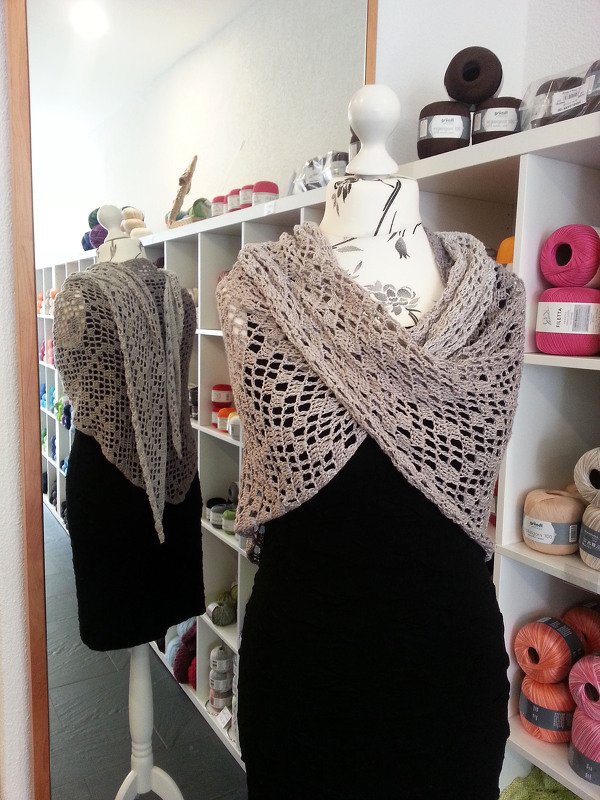 Hellbeiges gehäkeltes Dreieckstuch mit offenem Lochmuster, drapiert auf einer Schaufensterpuppe; Garnregal und Spiegelung im Hintergrund