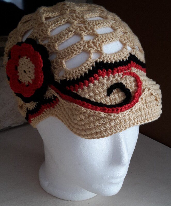 Beige gehäkelte Mütze mit roter Blume und schwarzen sowie roten Mustern auf einem Mannequinkopf