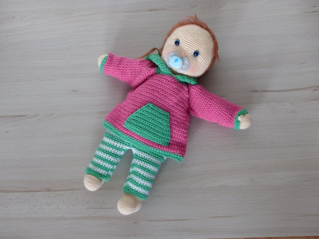 Crocheted amigurumi doll wearing a pink hoodie with a green pocket, green-and-white striped pants, and a blue pacifier.