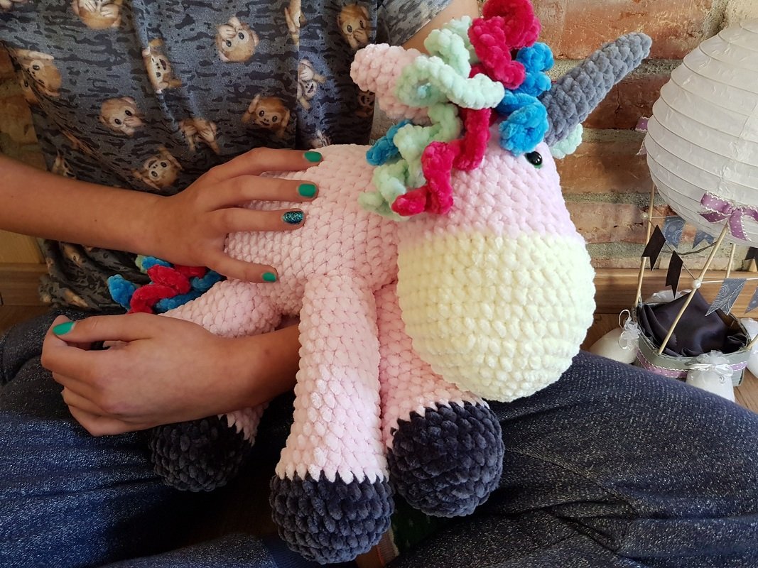Großes rosa gehäkeltes Einhorn-Amigurumi mit bunter Mähne und cremefarbener Schnauze, in Händen gehalten.
