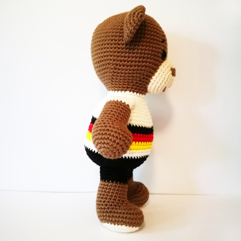 Gehäkelter brauner Amigurumi-Teddybär, Seitenansicht, weißes Shirt mit schwarzen, roten und gelben Streifen