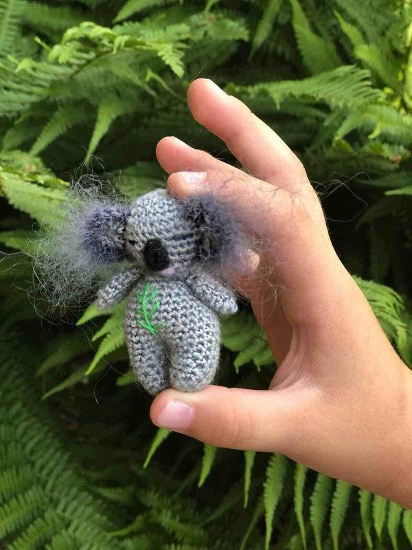 Hand hält winziges graues gehäkeltes Koala-Amigurumi vor Farnblättern.