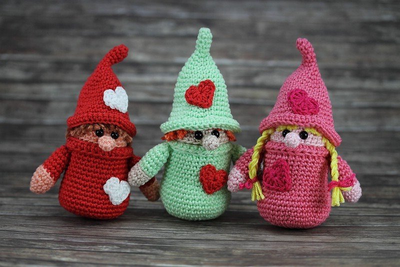 Drei gehäkelte Amigurumi-Wichtel in Rot, Mint und Pink mit Mützen und Herzmotiven vor Holz-Hintergrund