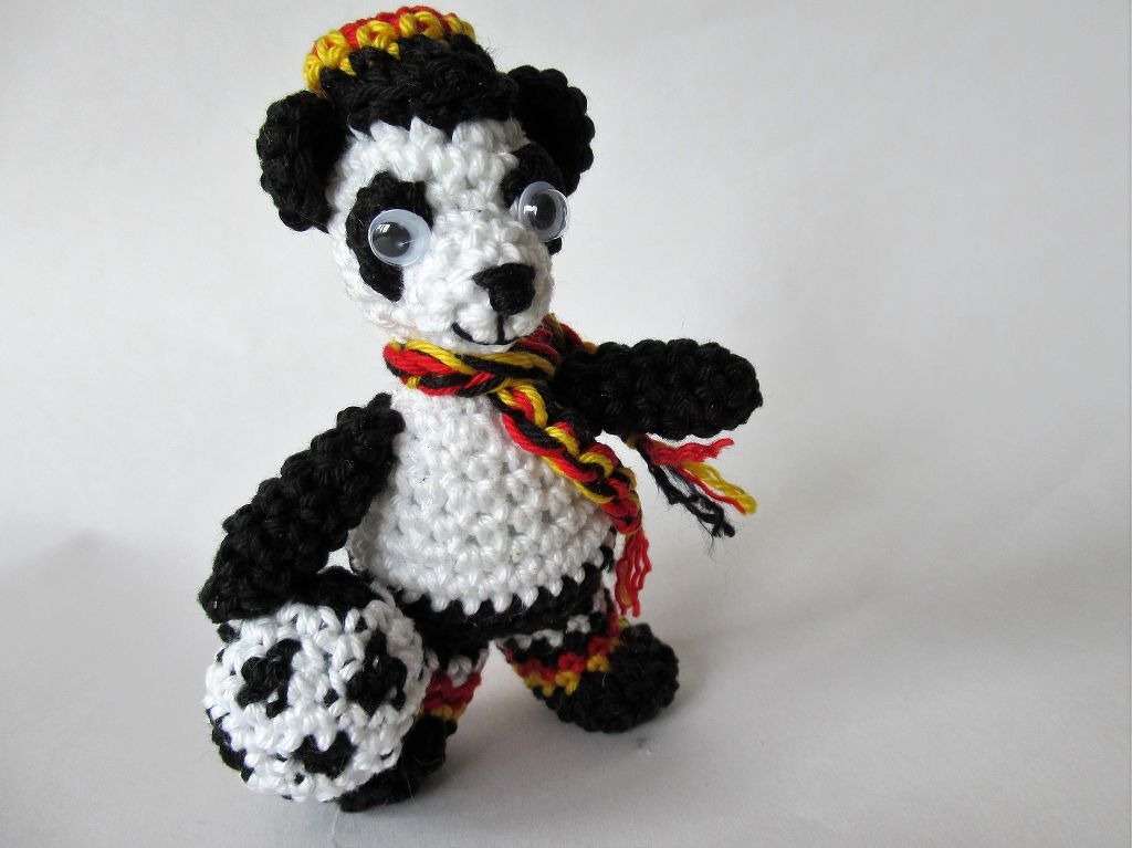 Gehäkelter Panda-Amigurumi hält einen kleinen Fußball und trägt Mütze sowie Schal in Schwarz
‑Rot‑Gelb.