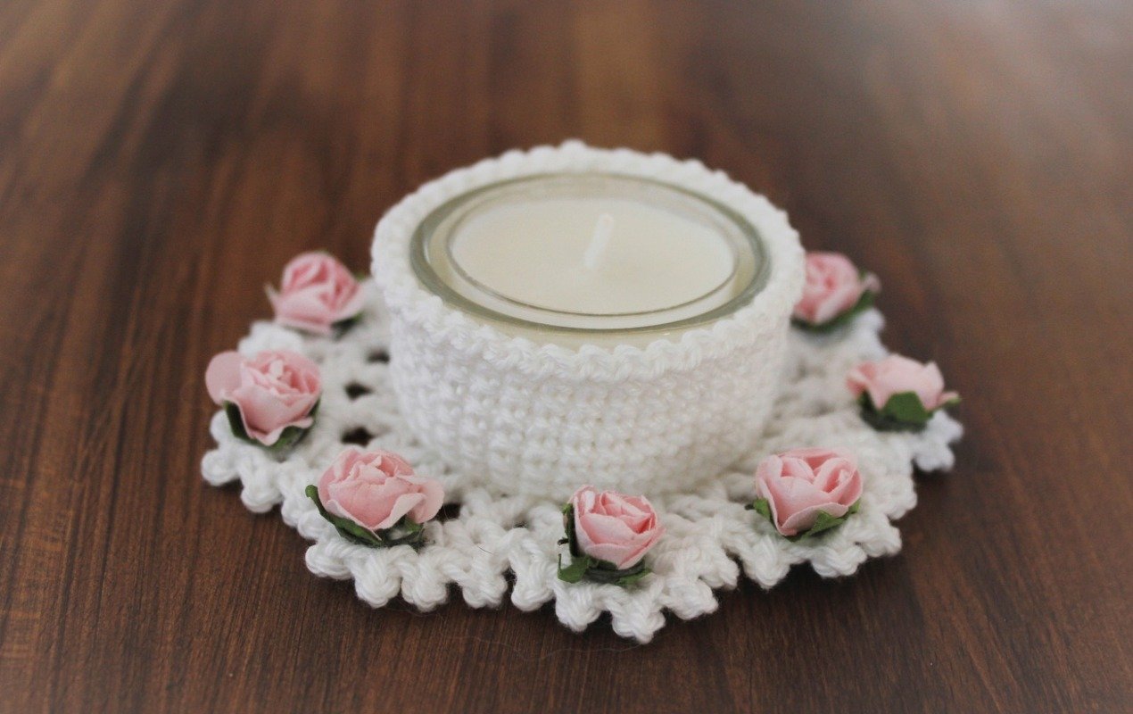 Weißer gehäkelter Teelichthalter mit rosa Rosen und Teelicht im Glas auf dunklem Holz.