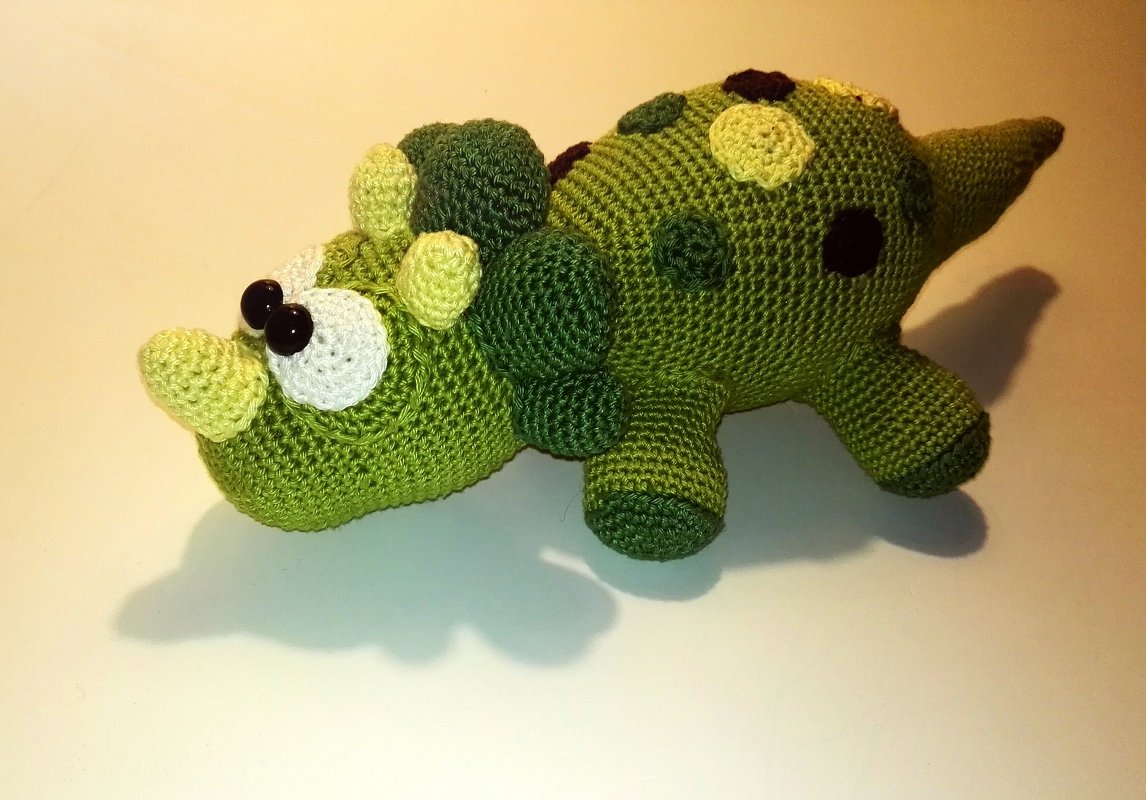 Gehäkelter grüner Amigurumi-Dinosaurier mit Hörnern und Punkten auf hellem Hintergrund