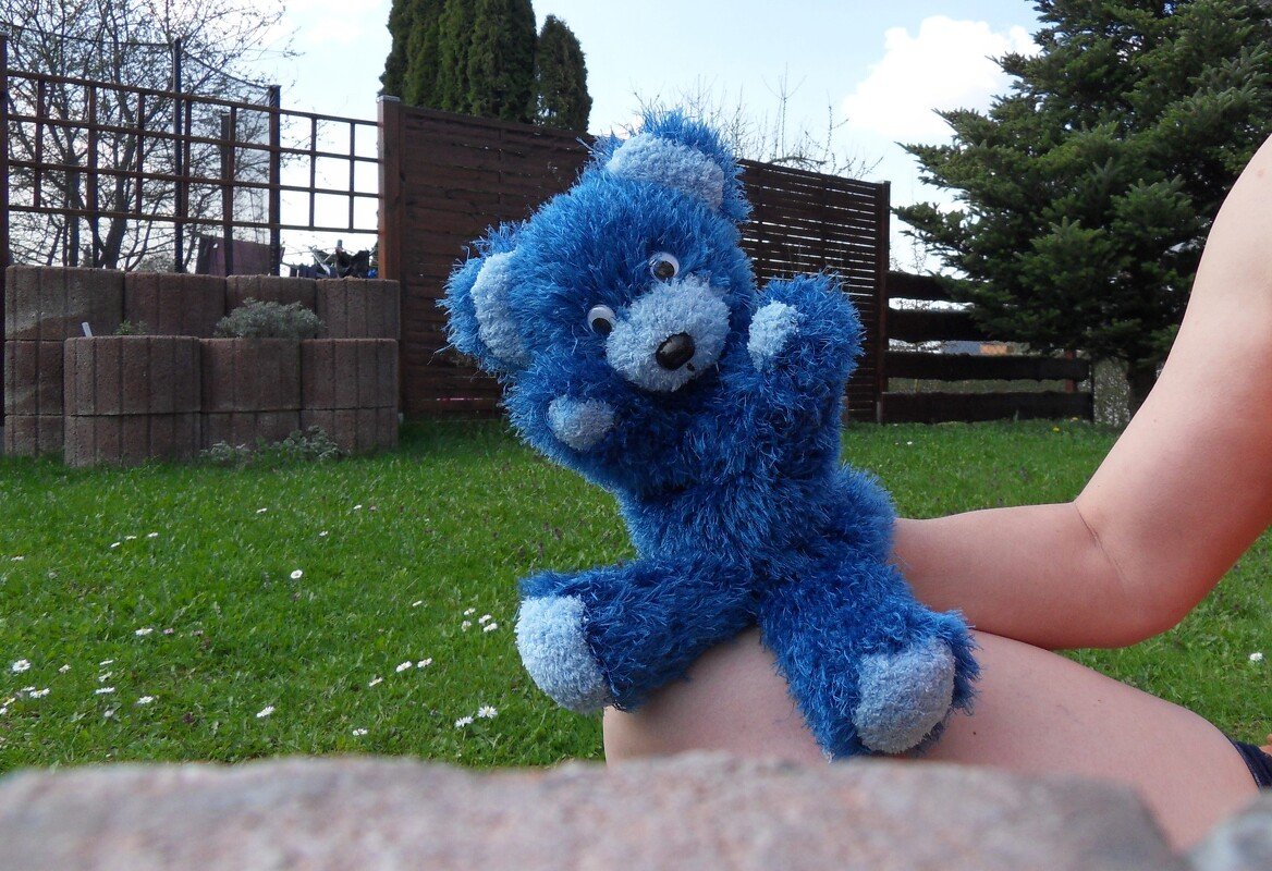 Blauer, flauschiger Teddybär mit hellblauen Akzenten und großen Augen auf dem Knie einer Person im Garten