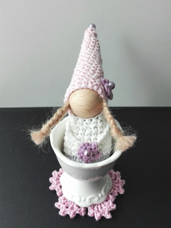 Gehäkelter Amigurumi-Wichtel mit Holzgesicht, rosa spitzer Mütze und geflochtenen Zöpfen im weißen Eierbecher auf rosa Häkeluntersetzer.