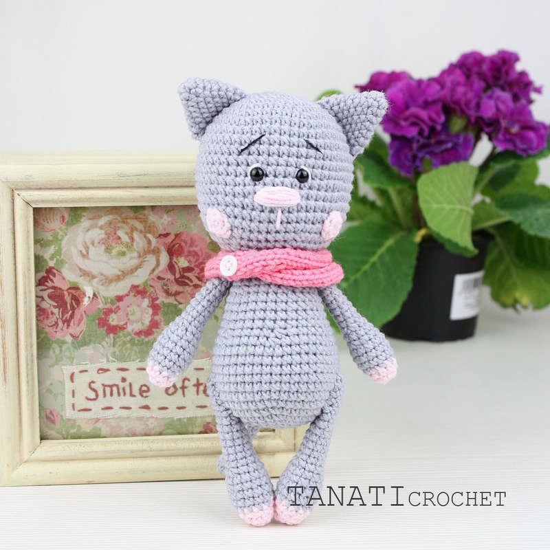 Gehäkelte graue Amigurumi-Katze mit rosa Schal vor Blumenbild
