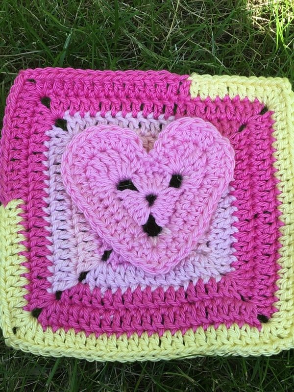 Gehäkeltes Granny Square in Pink und Gelb mit rosa 3D-Herz auf Gras.