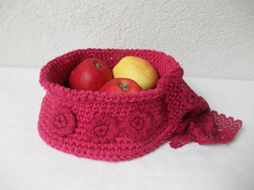 Small pink crochet storage basket with three floral appliqués and lace edge holding apples