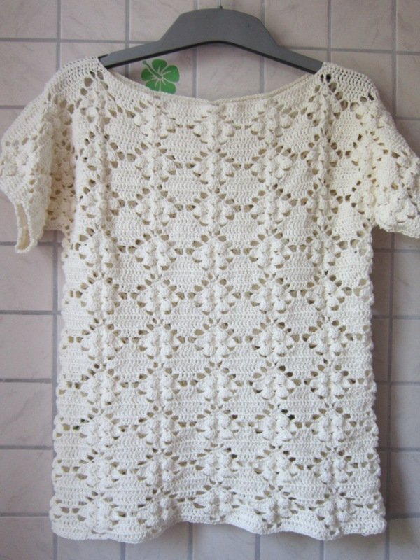 Cremefarbenes gehäkeltes Damen-Shirt mit kurzem Ärmel und dekorativem Loch- und Noppenmuster, an einem Kleiderbügel hängend.
