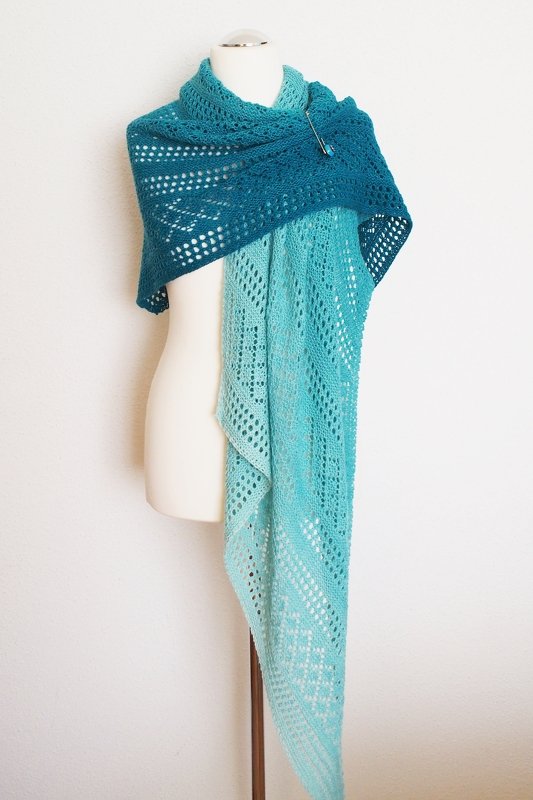 Long triangular knitted lace shawl in gradient teal to aqua draped on a white mannequin, secured with a shawl pin.