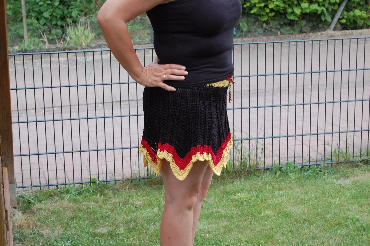 Person trägt einen gehäkelten schwarzen Minirock mit rot-gelbem, gewelltem Saum neben einem Drahtzaun
