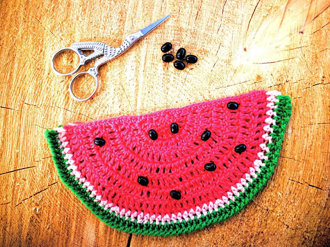 Gehäkelte Melonenscheiben-Applikation mit sichtbaren Maschen und schwarzen Perlen, liegend auf Holz neben kleiner Schere