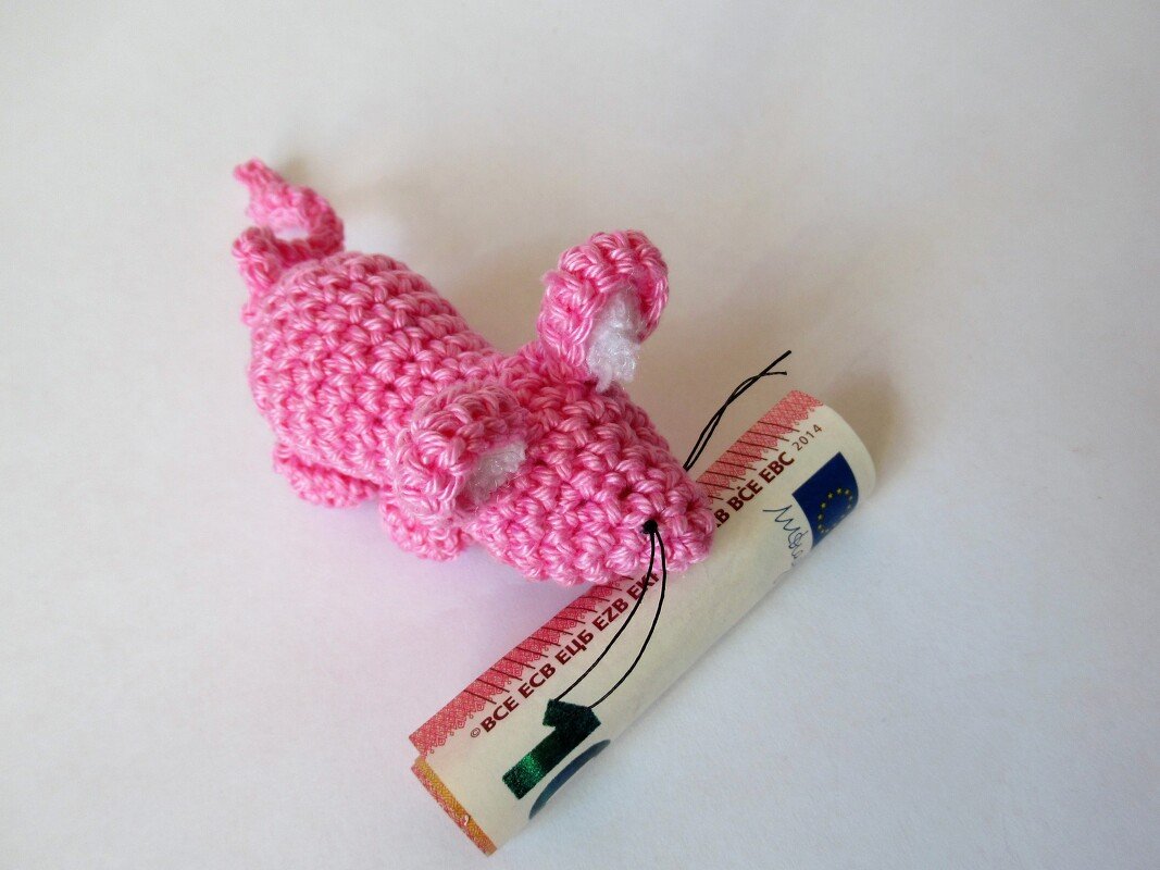Pinke gehäkelte Amigurumi‑Maus mit schwarzen Schnurrhaaren neben einem zusammengerollten Geldschein