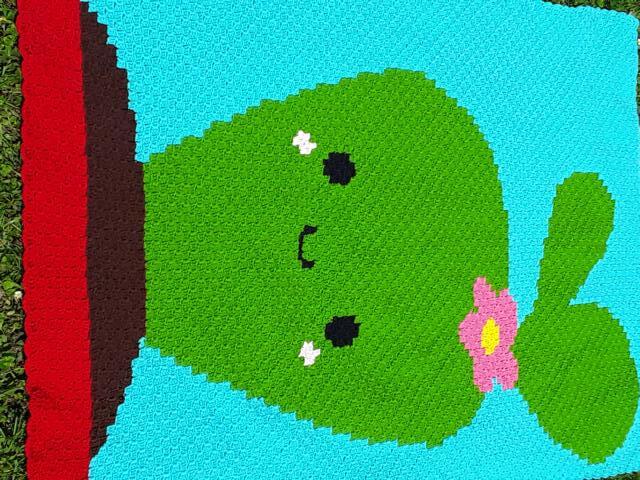 Gehäkelte Babydecke mit grünem Kaktus, rosa Blüte und braunem Topf auf hellblauem Hintergrund mit rotem Rand.