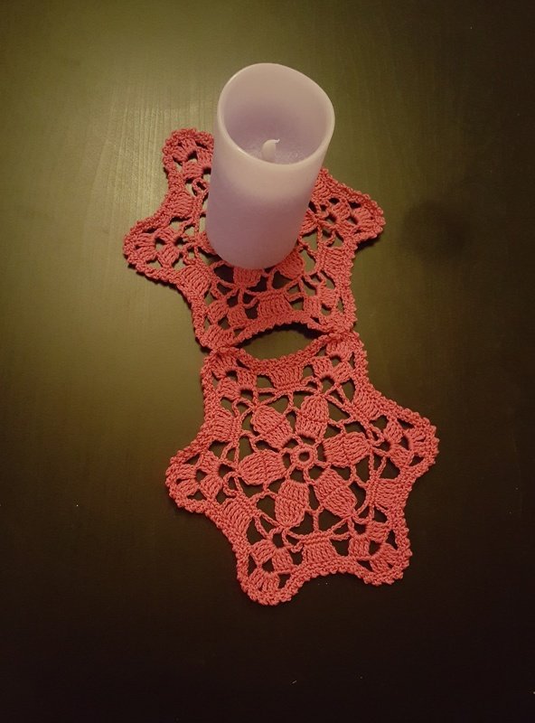 Zwei rosa gehäkelte sternförmige Deckchen auf dunklem Tisch, eines mit hellvioletter Kerze.