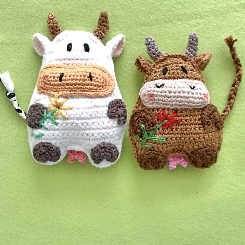 Zwei gehäkelte Kuh-Amigurumi-Figuren (weiß und braun) mit bestickten Blumen auf grünem Stoff