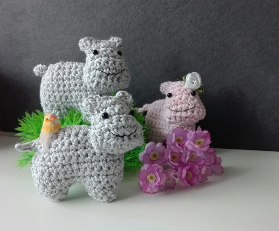 Drei gehäkelte Nilpferd-Amigurumi, zwei grau und eins rosa, mit schwarzen Augen und dekorativen Blumen