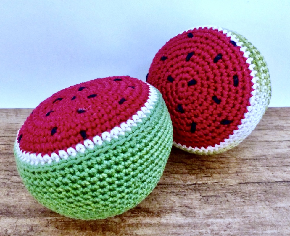 Zwei gehäkelte Wassermelonenhälften in Rot, Weiß und Grün mit schwarzen Kernen auf Holzuntergrund.