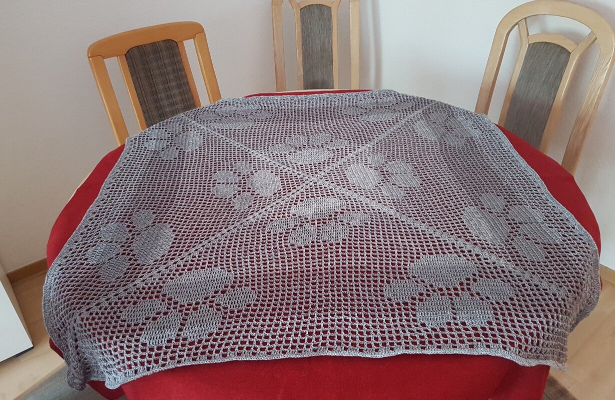 Quadratisches Filetdeckchen mit wiederkehrenden Blütenmustern auf rundem Tisch mit rotem Tischtuch