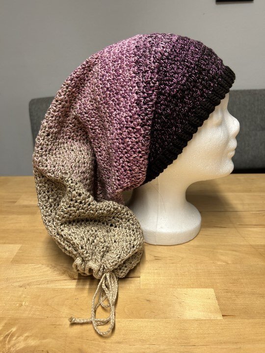 Seitenansicht eines gehäkelten Scoodies in Beige‑Rosa‑Violett mit geripptem Rand und Kordel auf weißem Schaufensterkopf