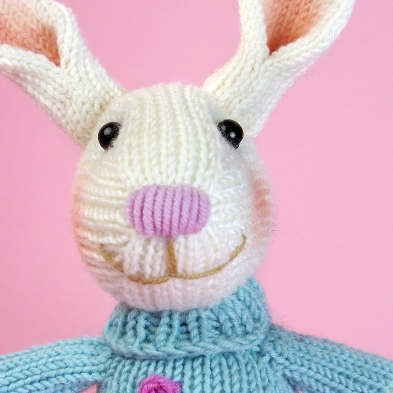 Close-up of a white knitted bunny with a pink nose and blue sweater on a pink background.