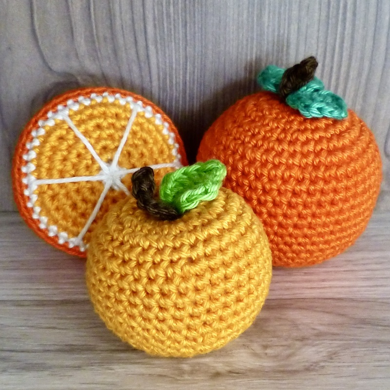 Nahaufnahme: zwei gehäkelte Orangen und eine Orangenscheibe in Orange- und Gelbtönen auf hellem Holz.