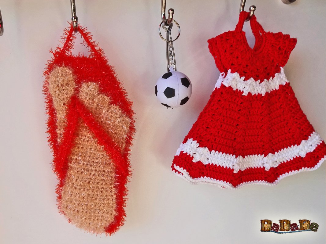 Zwei gehäkelte Anhänger an Haken: rotes Mini-Kleid mit weißen Streifen und beige-rote flauschige Badelatsche; dazwischen kleiner Fußball‑Schlüsselanhänger.