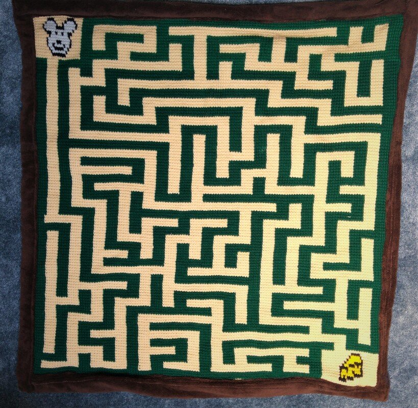 Gehäkelte Decke mit labyrinthartigem Pixeldesign in Grün und Creme, dunkelbrauner Rand, mit Pixelmotiven von Maus und Käse in den Ecken