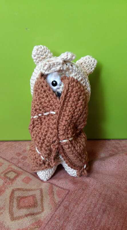 Gehäkelte Amigurumi-Figur in beigefarbener Kapuze mit Ohren, im braunen Wickelmantel ein Auge blickt hervor