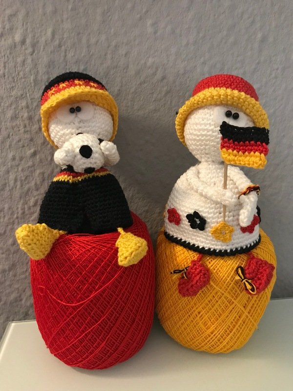 Zwei gehäkelte Amigurumi-Puppen in schwarz-rot-gold auf roten und gelben Garnknäueln, eine mit kleinem Fußball, eine mit Mini-Fahne