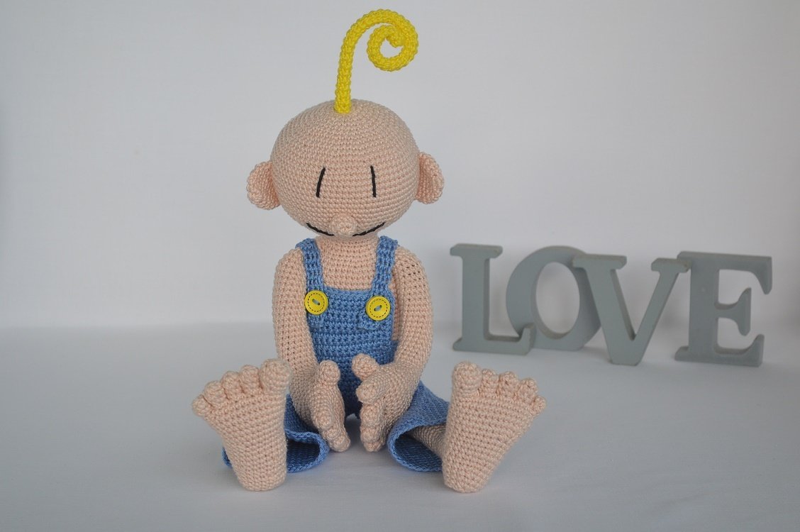 Amigurumi baby doll in blue overalls with yellow curl, seated next to grey LOVE letters on a white background.