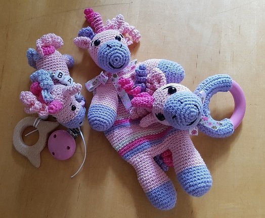 Gehäkeltes Baby-Set mit Einhorn-Motiv: Ein Schmusetuch, eine Schnullerkette mit Buchstaben und ein Greifling in Rosa, Lila und Hellblau auf einem Holztisch