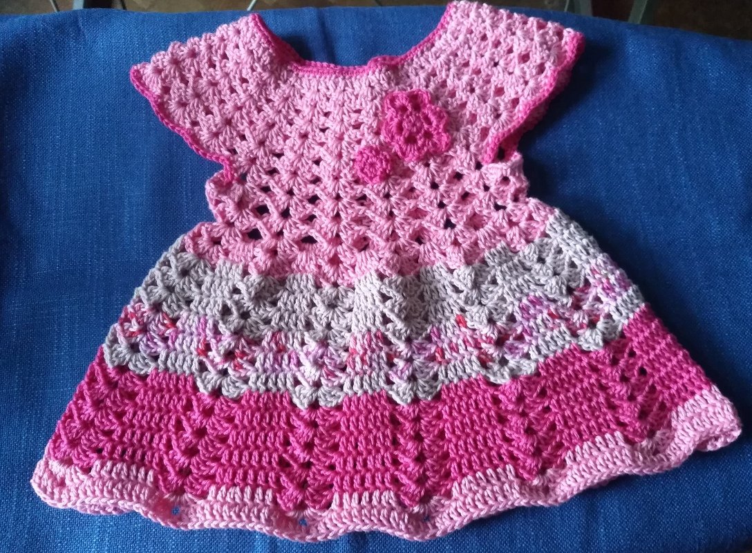 Gehäkeltes Babykleid in drei Rosa- und Pinktönen mit verschiedenen Musterbereichen und applizierten Häkelblumen, liegend auf blauem Stoff.