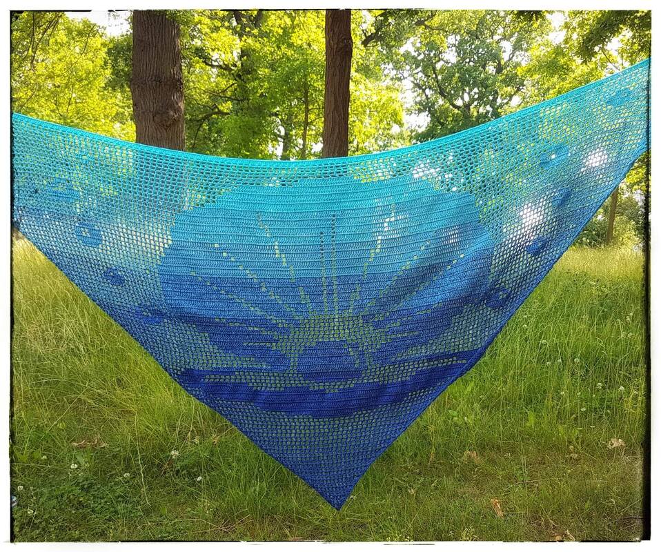 Türkis-blaues gehäkeltes Dreieckstuch mit Sonnenstrahlen-Motiv, im Grünen aufgehängt