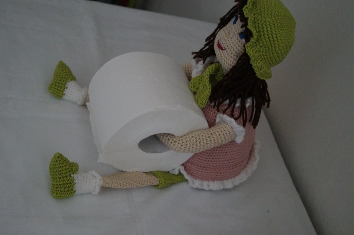 Handgehäkelte Amigurumi-Puppe in rosa Kleid und grüner Mütze, die eine Toilettenpapierrolle hält.