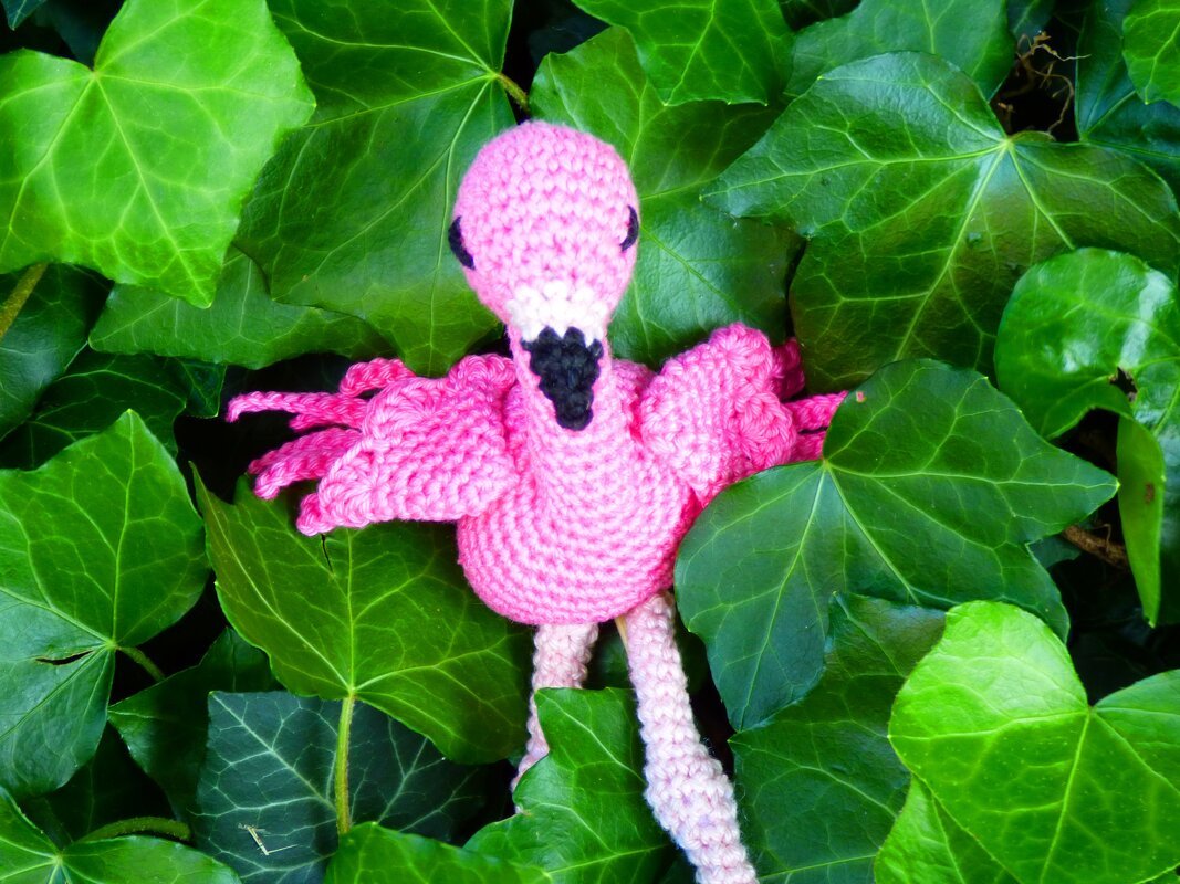 Nahaufnahme eines gehäkelten pinken Flamingos mit sichtbarer Maschenstruktur auf grünem Efeuhintergrund