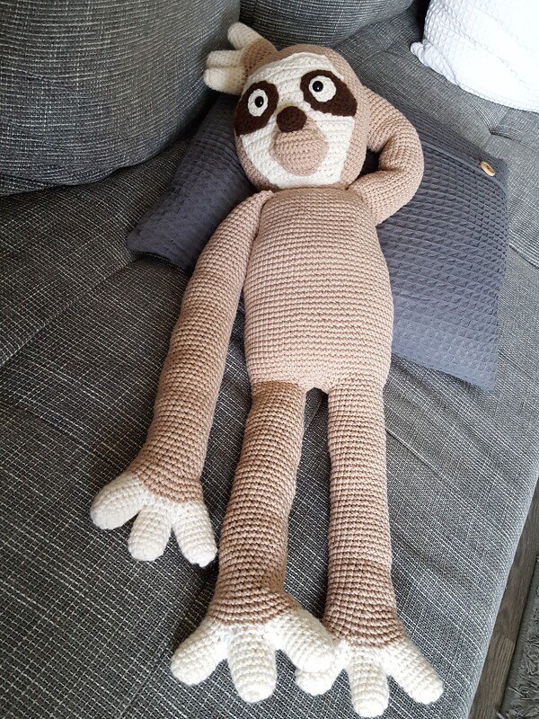 Gehäkeltes Amigurumi-Faultier (beige/creme) mit langen Armen, auf grauem Sofa liegend