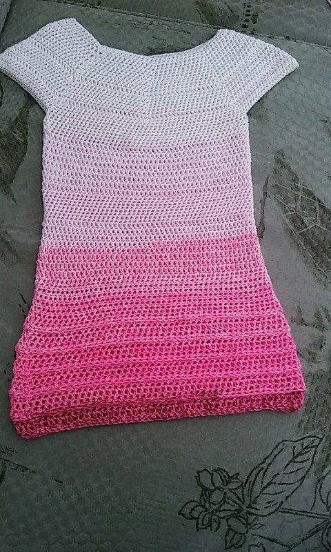 Gehäkeltes Kinderkleid mit Rosa-Ombré, kurzen Ärmeln und rundem Ausschnitt, liegend auf dunklem Untergrund.