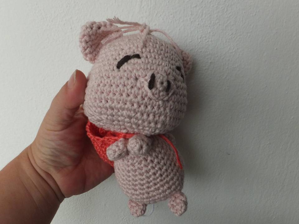 Hand hält ein gehäkeltes rosa Amigurumi‑Schwein mit rotem Halstuch vor heller Wand