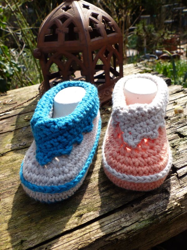 Zwei handgehäkelte Babyschuhe in Türkis‑Grau und Rosa‑Grau auf verwittertem Holz mit Laterne im Hintergrund.