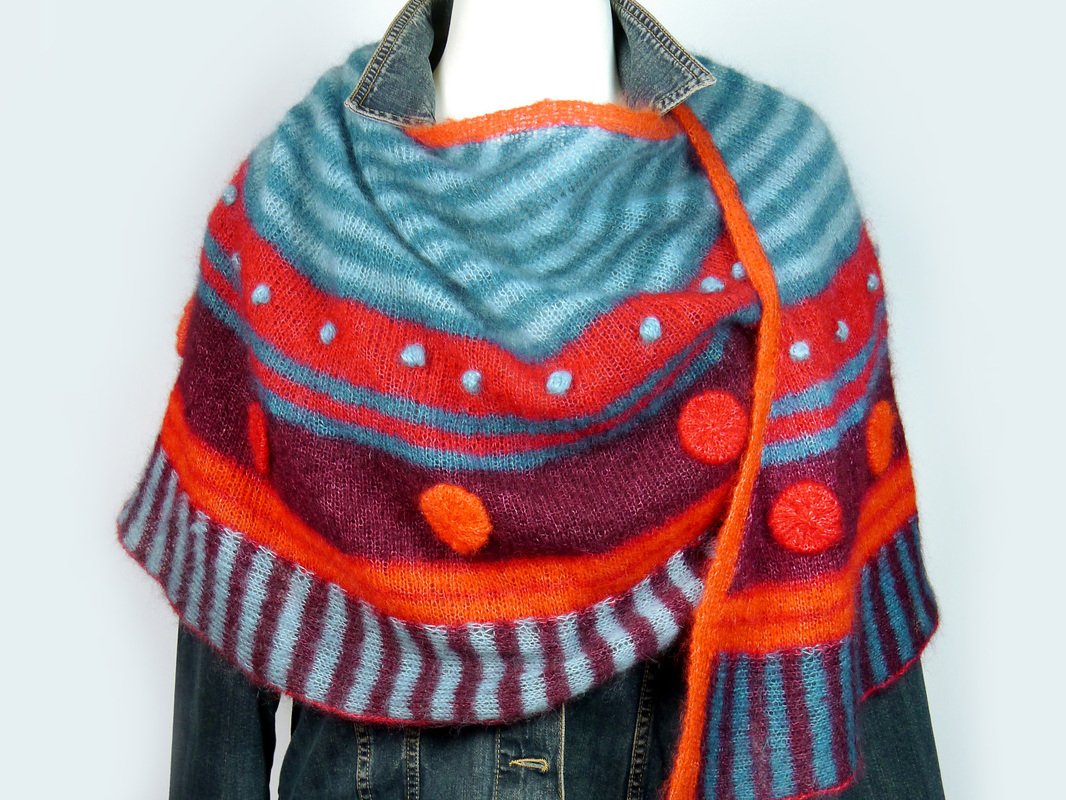 Crescent shawl in teal, plum and orange with stripes and bobbles draped over a denim-collared mannequin